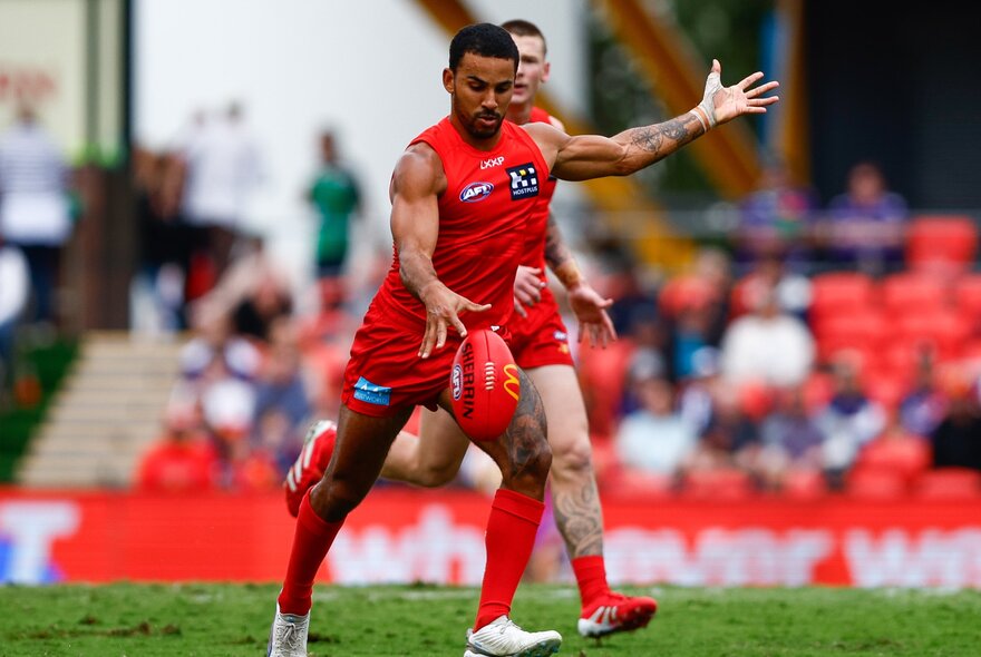 A Gold Coast Suns AFL football player on the field kicking a red football, blurred spectators in the background.