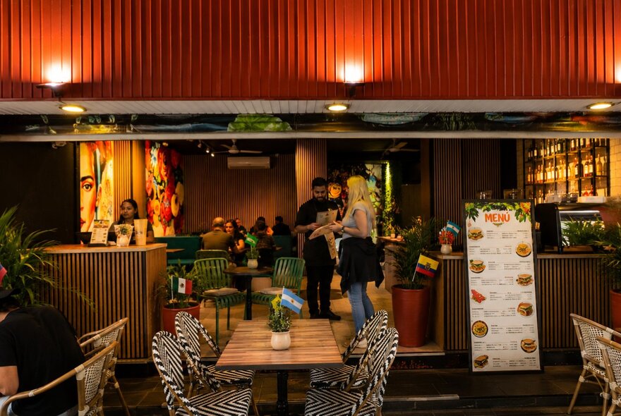 Outdoor or patio dining area of a restaurant at night, with several small tables, wooden accents, and a large menu board standing near the entrance.