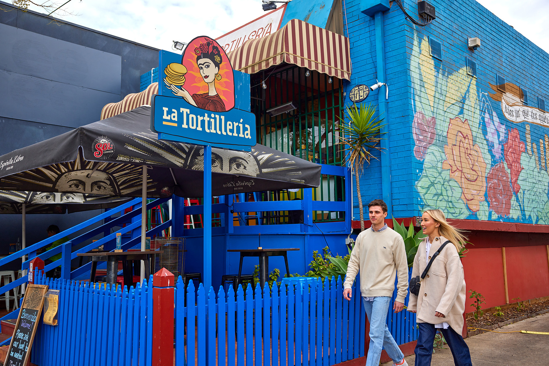 Two people walking into a blue restaurant with mural and picket fence.