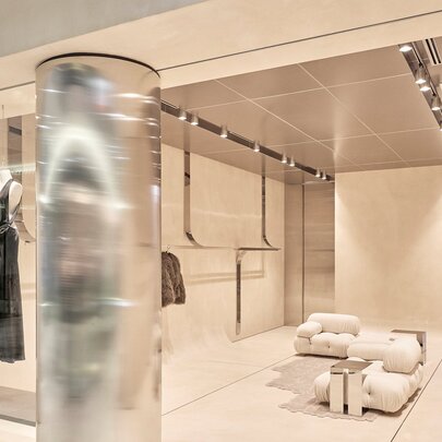 The exterior of a luxury fashion store with minimalist decor and a dress in the window. A metallic column partially blocks the view. 