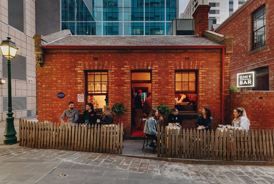 Red brick exterior of Little Lon Distilling showing patrons sitting outside, bluestone paving in front and city skyscrapers in the background.