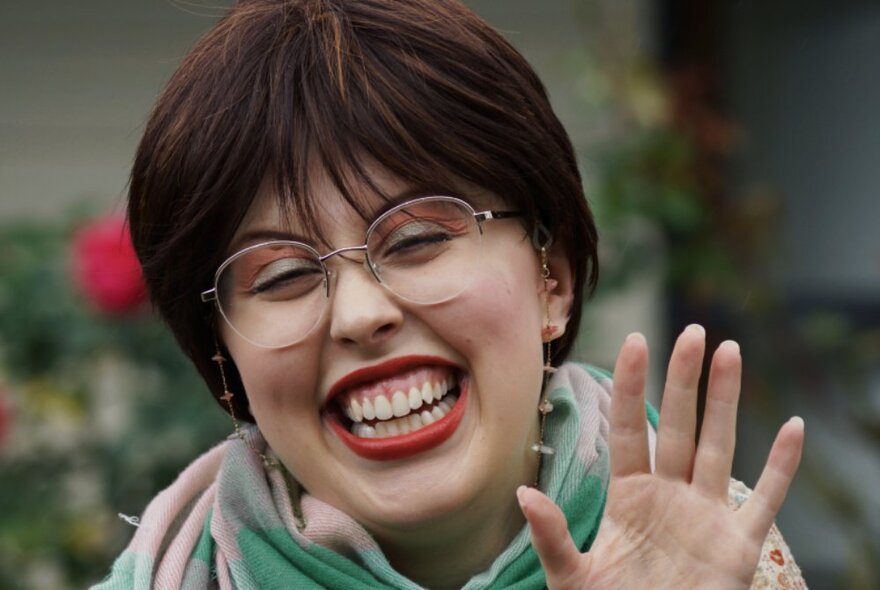 Performer with short hair and glasses grinning widely and raising a hand.