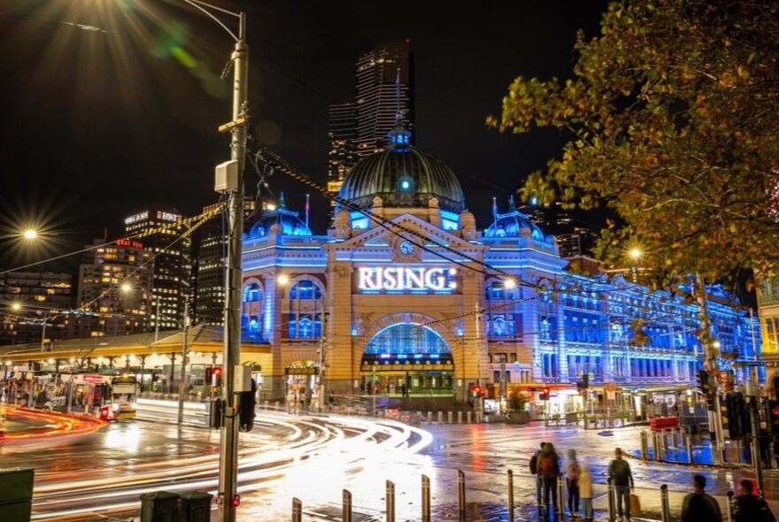 Flinders Street Station lit blue at night with a large sign that reads RISING.