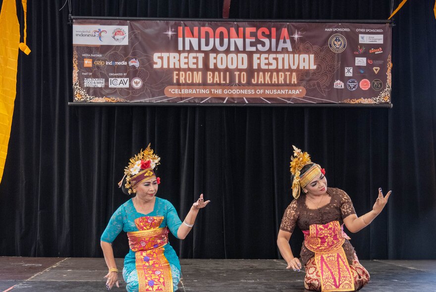 Two Indonesian dancers performing on a stage with a brown banner and black curtain behind them.