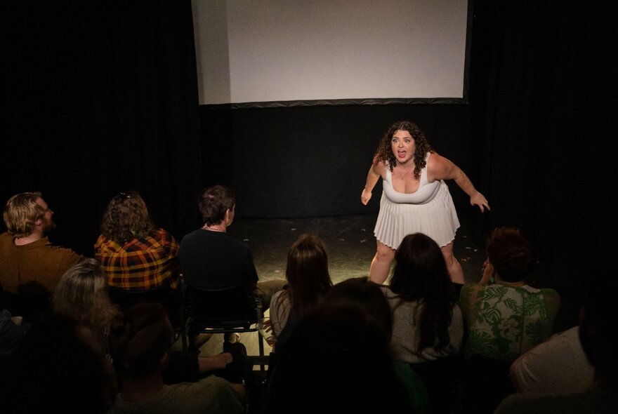 A woman in a white dress stands on a dimly lit stage, performing comedy in front of a seated audience whose backs are mostly visible.