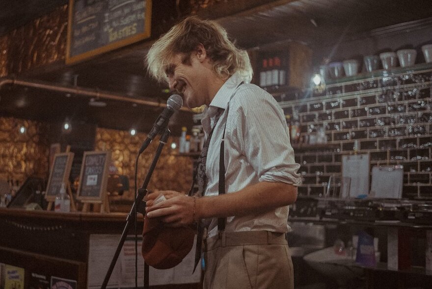 Man standing at a microphone stand laughing in a brick-lined bar venue.