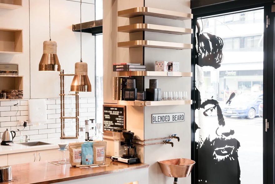 Cafe interior with white-tiled kitchen area, copper shelves and lighting, and artwork of bearded man on shopfront glass.