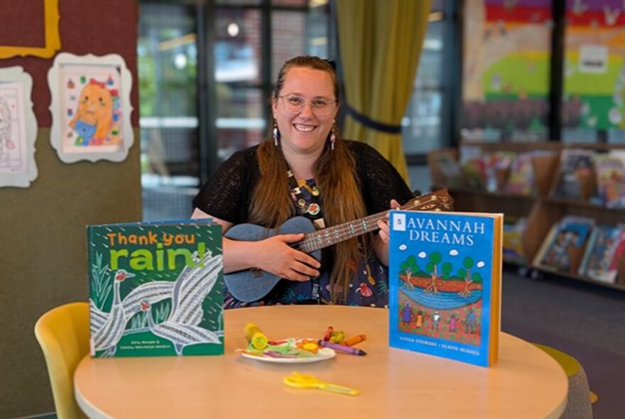 A smiling adult holding a ukulele seated at a low table, with two picture books resting on the table, in a library setting.