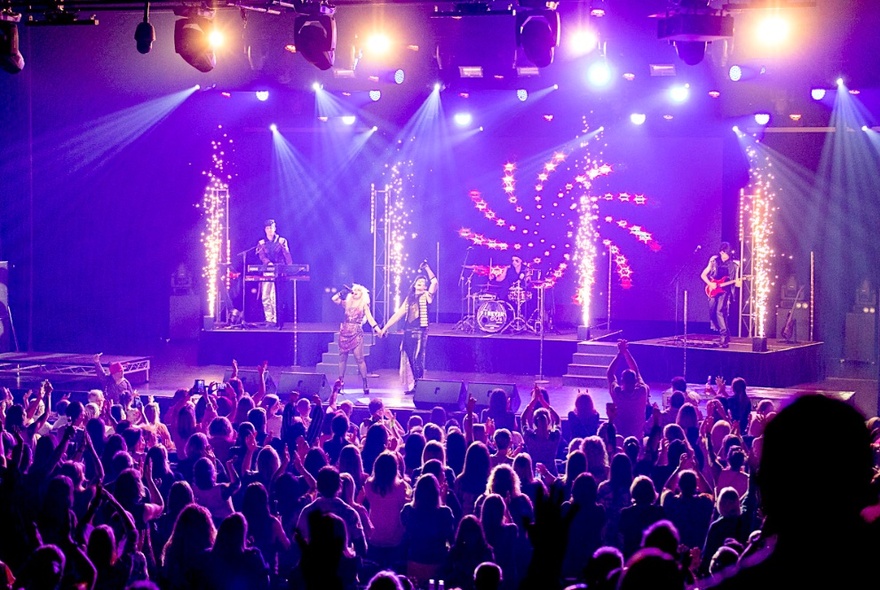Five 80s-style rockers on stage with theatrical flames and strobe lighting, in front of a crowd.