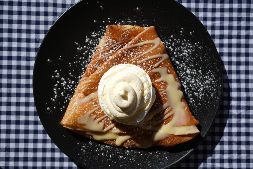 A classic triangular shaped French crepe with a whipped cream on a black plate on a black gingham tablecloth.