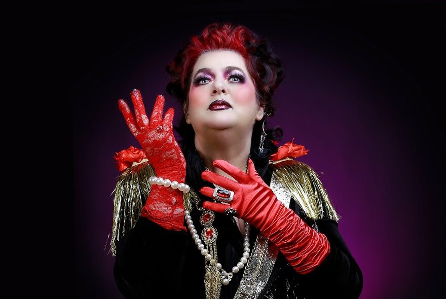 Performer wearing bold makeup, red gloves, gold cape and jewellery, gesturing with her hands and looking skywards.