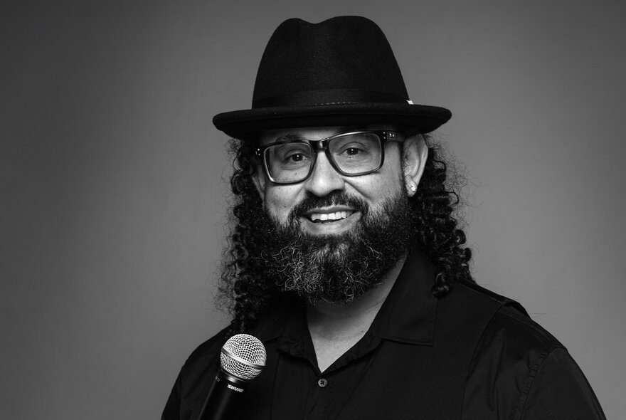 A man with a beard and curly hair, wearing a black fedora-style hat, yellow-tinted glasses, and a black shirt, holding a microphone and smiling at the camera; black and white photograph.