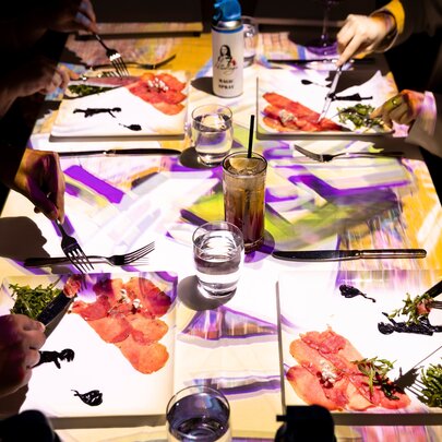 Diners seated at a table and eating from plates that have designs projected onto them from above, as part of an immersive visual art and food experience.