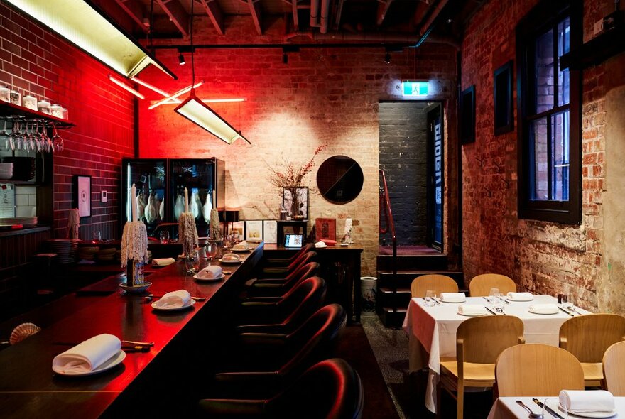 The moody interior of a Chinese restaurant, with exposed brick walls, tables with white tablecloths on the right and a long dark bar with barstools on the left, a red light fitting in the background.