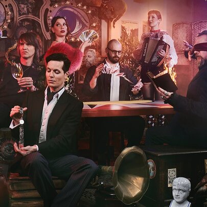 A group of people, including a blindfolded man, seated around a table in a dimly lit, ornate room for a magic show.