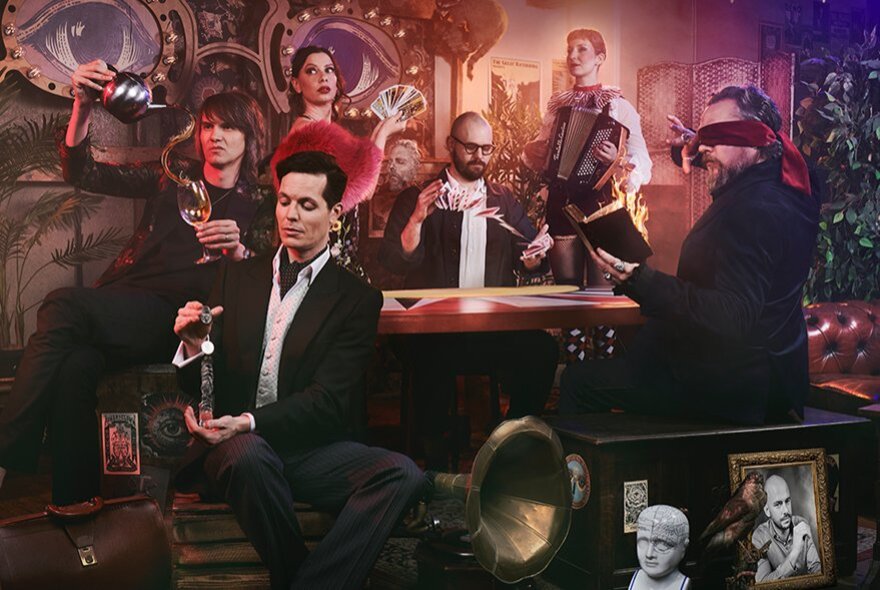 A group of people, including a blindfolded man, seated around a table in a dimly lit, ornate room for a magic show.