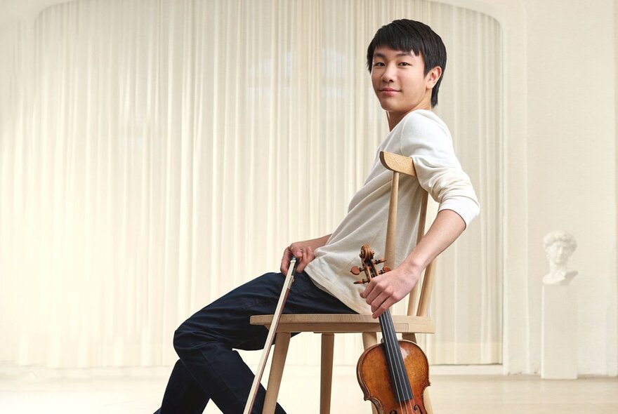 Classical musician Christian Li leaning back in a chair, holding a violin and bow.