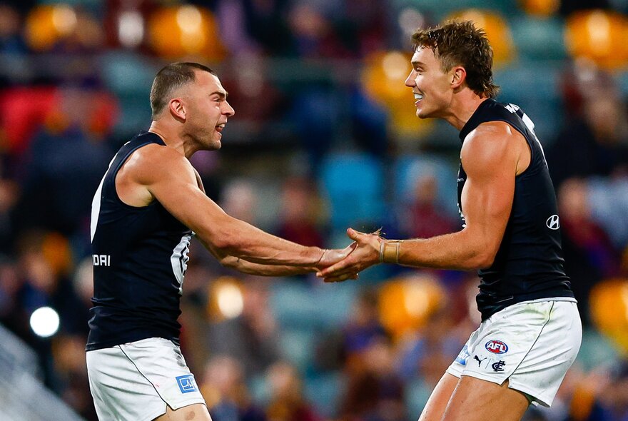 Two Carlton AFL players celebrating on the field after having scored, they face each other and are high-fiving their hands, with blurred fans in the stadium in the background.