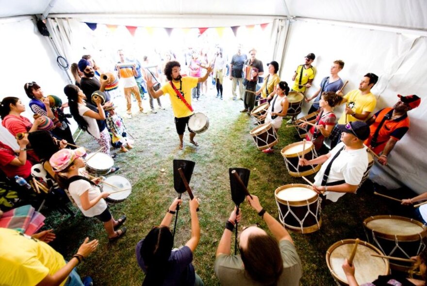 A group of people learning Afro-Brazilian percussion in a marquee, led by a man in a yellow t-shirt.