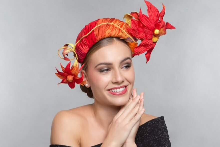 A smiling fashion model wearing a red and orange headpiece that features soft sculpted flowers.  
