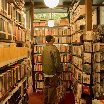 A person with short dark hair stands with their back to the camera, looking at shelves in a narrow, crowded bookstore; the room is filled from floor to ceiling with books, creating a cozy atmosphere. 