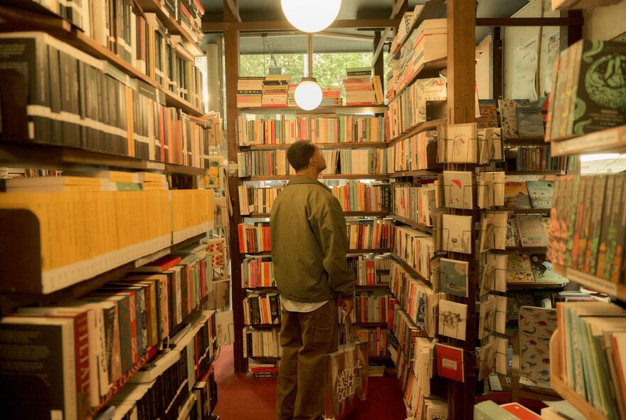 A person with short dark hair stands with their back to the camera, looking at shelves in a narrow, crowded bookstore; the room is filled from floor to ceiling with books, creating a cozy atmosphere. 
