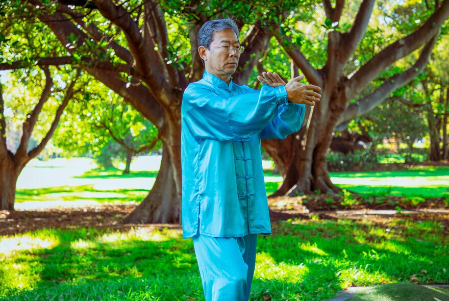 A man in a blue silk traditional Chinese outfit doing Tai Chi in a park, large trees behind him.