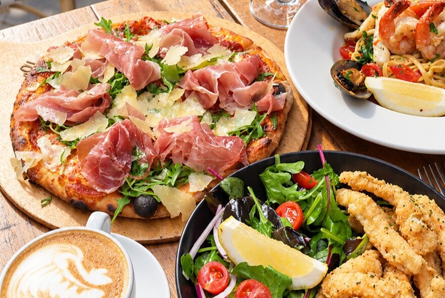 A top-down view of a dining table with a prosciutto and rocket pizza, a seafood pasta dish, a side salad, fried appetizers, and a cup of coffee.
