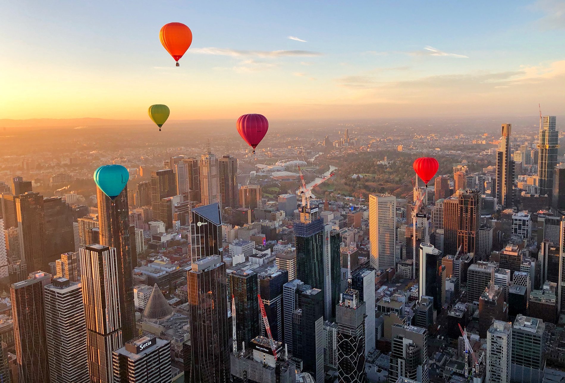 Colourful hot air balloons flying over Melbourne city buildings at sunrise.