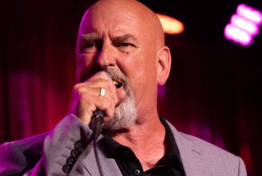 A middle-aged man with no hair and a beard, singing into a microphone on a stage.