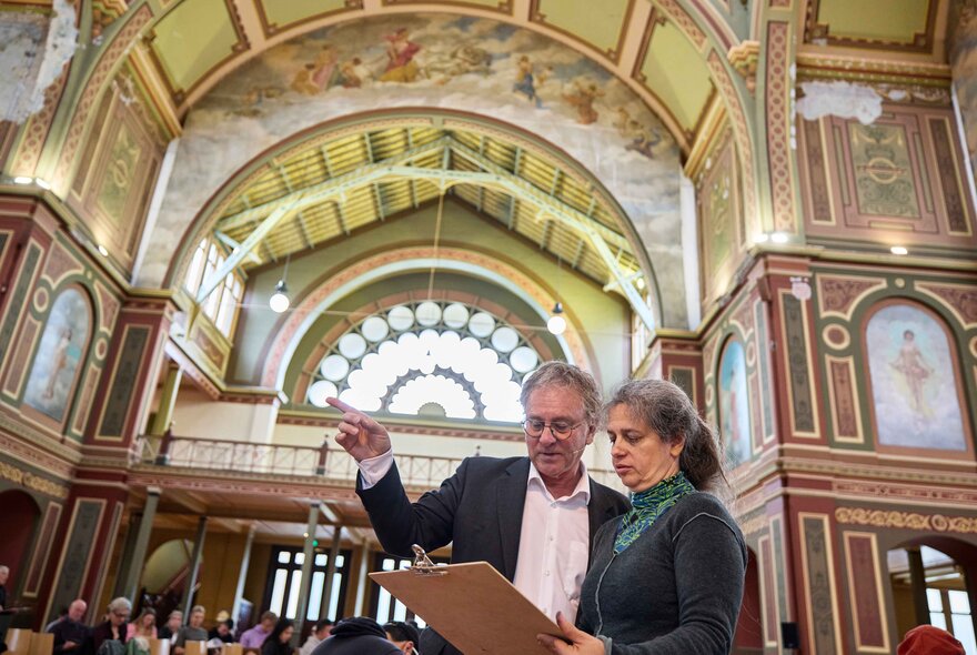 Two people review a sketch on a clipboard inside the ornate, mural-filled interior of the Royal Exhibition Building in Melbourne.