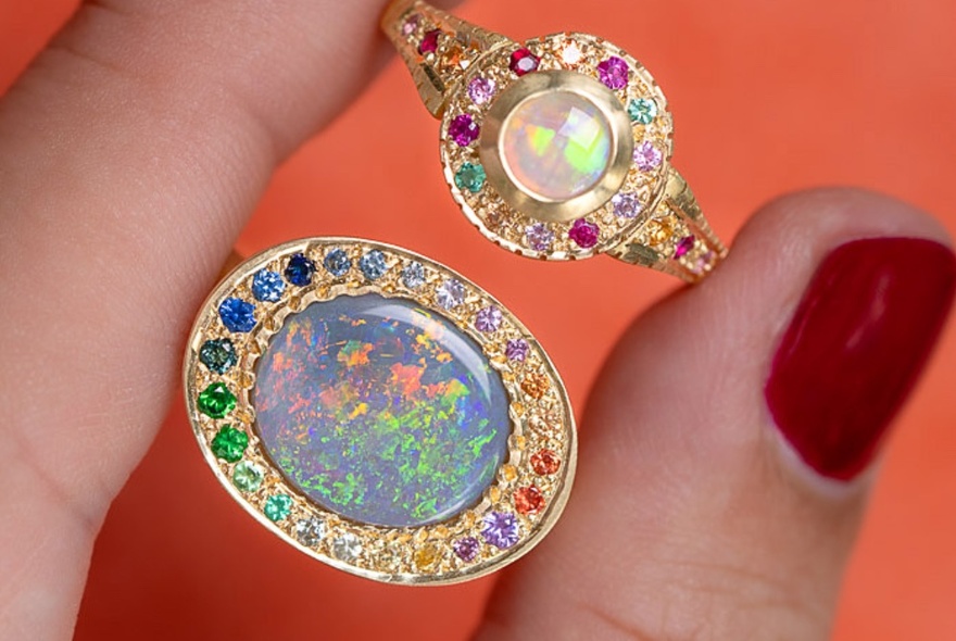 A hand holding two rings, one set with a multi-coloured opal and the other with a central pearl, both rings surrounded by smaller coloured gemstones. 