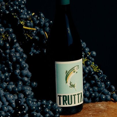 A bottle of Trutta wine (a trout on the label) nestled into a pile of dark wine grapes. 