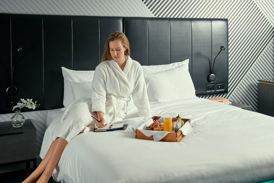 A woman in a white dressing robe writing in a notebook, while seated on a hotel bed, a tray of breakfast food on the bed beside her.