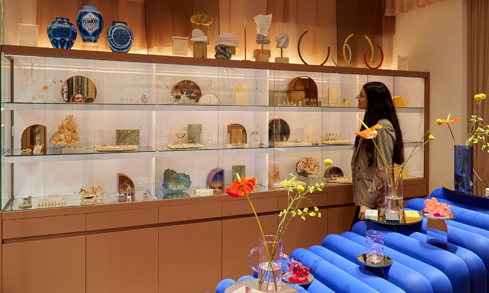 A woman looking at a jewellery cabinet in a store with a blue couch and floral displays.