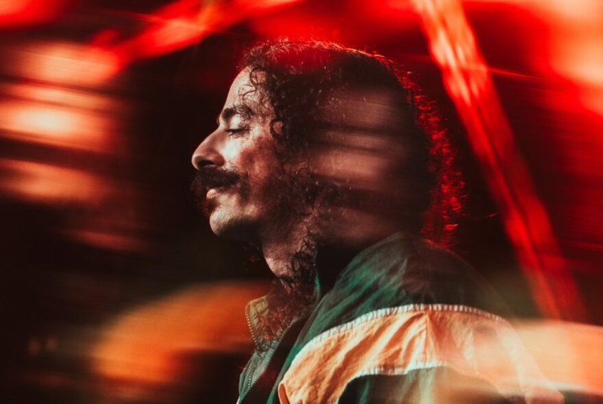 Singer Anees, a man standing in profile, with curly hair and a moustache, a red filter over the image.