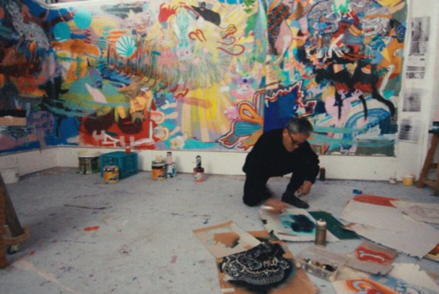 An artist painting in a workshop with brightly coloured murals on the wall, artist's materials on the floor.