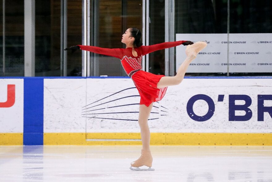 A female figure skater performing on ice in a red dress with her arms out and a leg in the air behind her. 