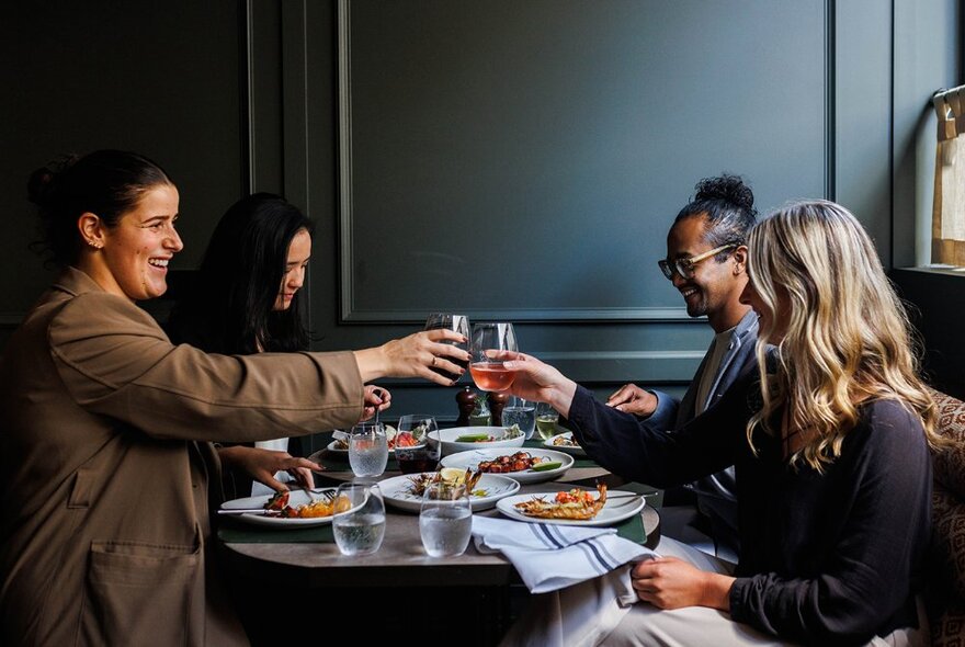 A group of four people enjoying dinner at a restaurant table, with plates of food in front of them and two holding up wine glasses.