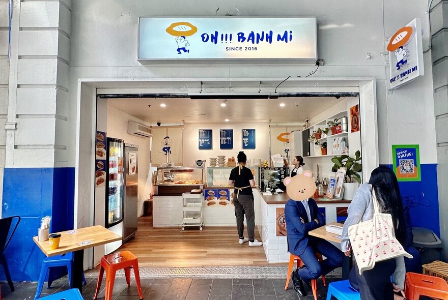 People seated at an outdoor cafe table, with view into ordering counter and kitchen.