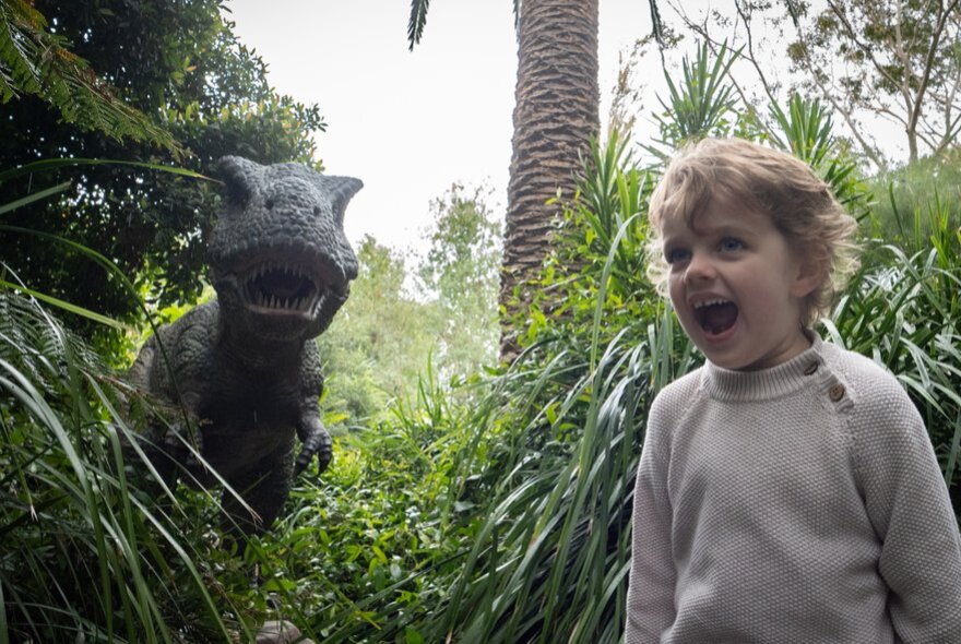 A large dinosaur model with its mouth open in a park setting with a child, also open-mouthed, in the foreground. 