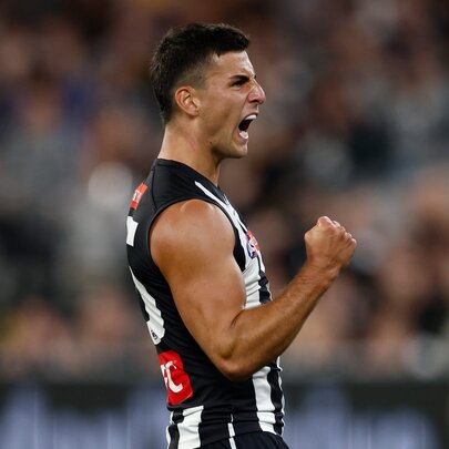 A Collingwood AFL football player with his fist clenched and pumped during a match, a blurred stadium crowd in the background.