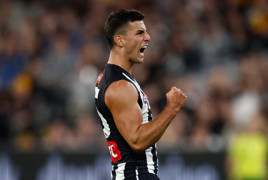A Collingwood AFL football player with his fist clenched and pumped during a match, a blurred stadium crowd in the background.