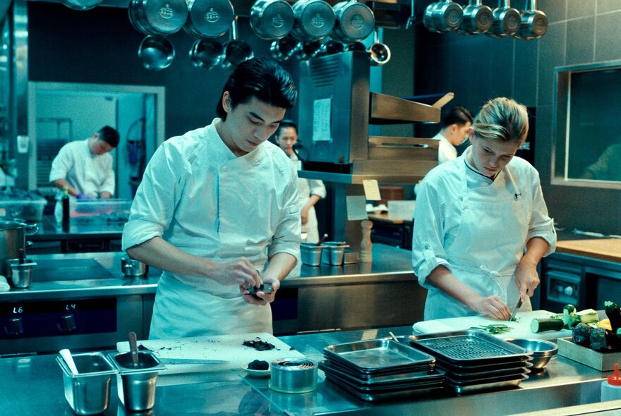 A still from the film Redress showing a man and a woman in chef uniforms preparing food at a stainless steel counter in a professional kitchen.