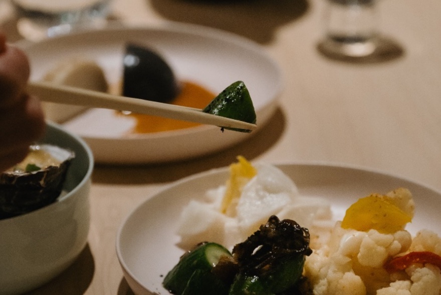 Chopsticks selecting a small pickled vegetable piece from a plate of food on a table.