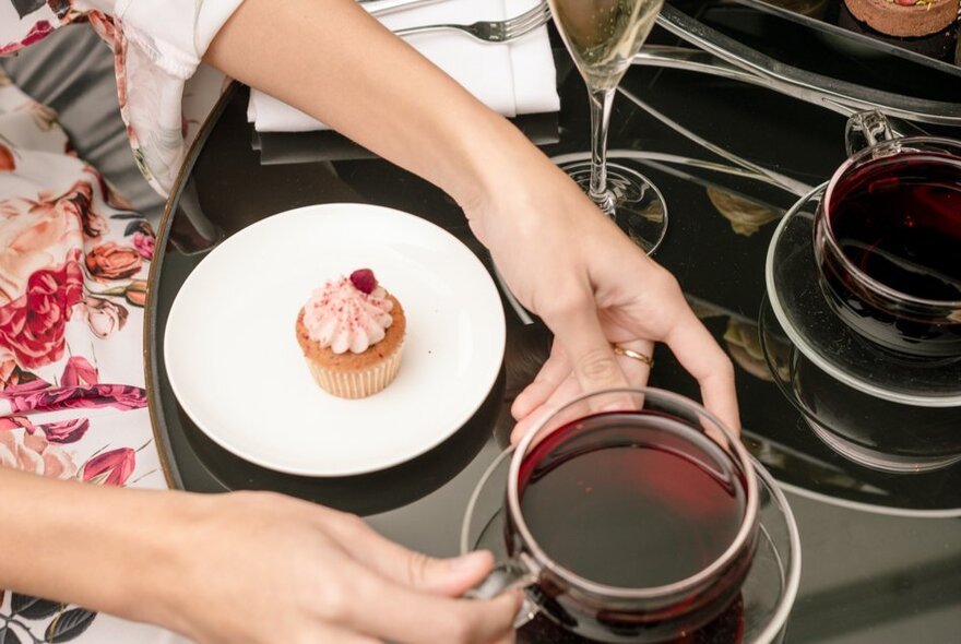A woman's hands picking up a glass teacup and saucer; a small white plate next to it with a cupcake on it.