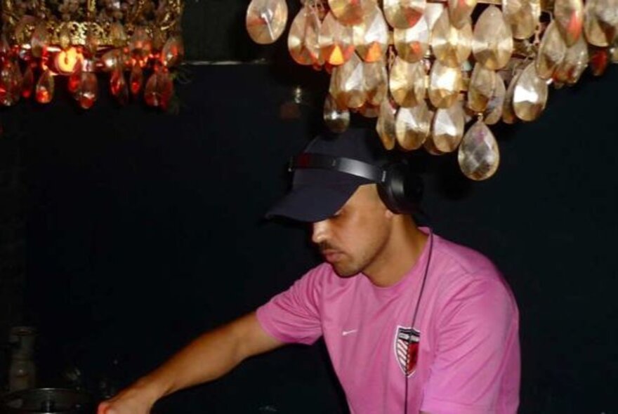 A man wearing a pink t-shirt, black cap and headphones at a DJ mixing desk.