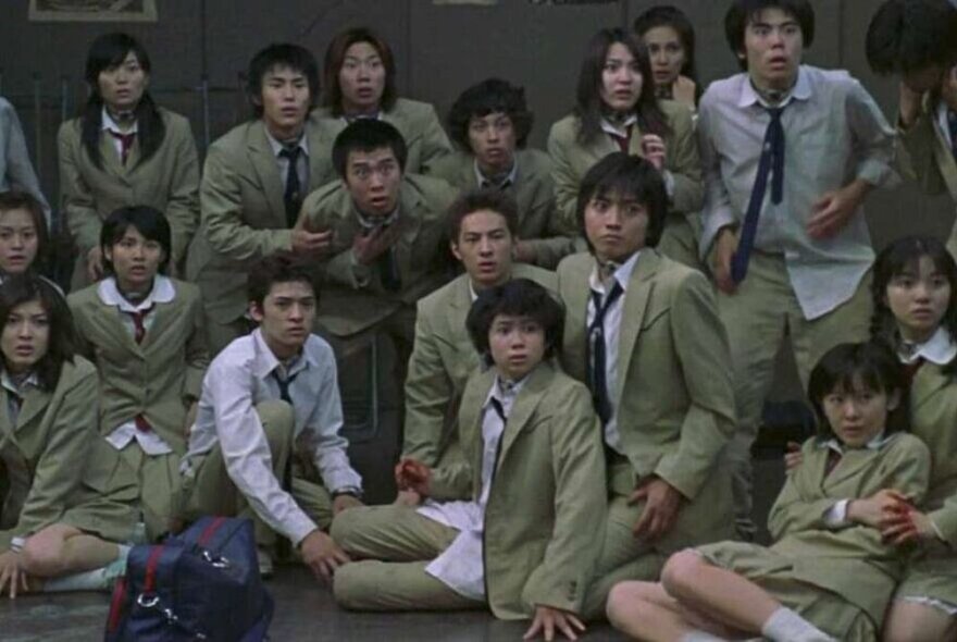 Film still of a large group of school students seated, wearing beige uniforms, looking terrified.