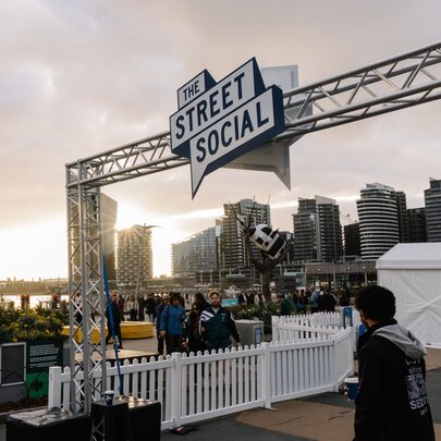 An outdoor space at Harbour Esplanade Docklands, featuring a large overhead hanging sign that reads THE STREET SOCIAL, a white picket fence, DJ equipment and a view of the harbour and city buildings.