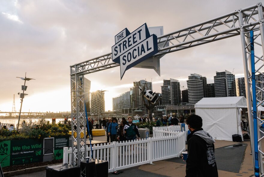 An outdoor space at Harbour Esplanade Docklands, featuring a large overhead hanging sign that reads THE STREET SOCIAL, a white picket fence, DJ equipment and a view of the harbour and city buildings.